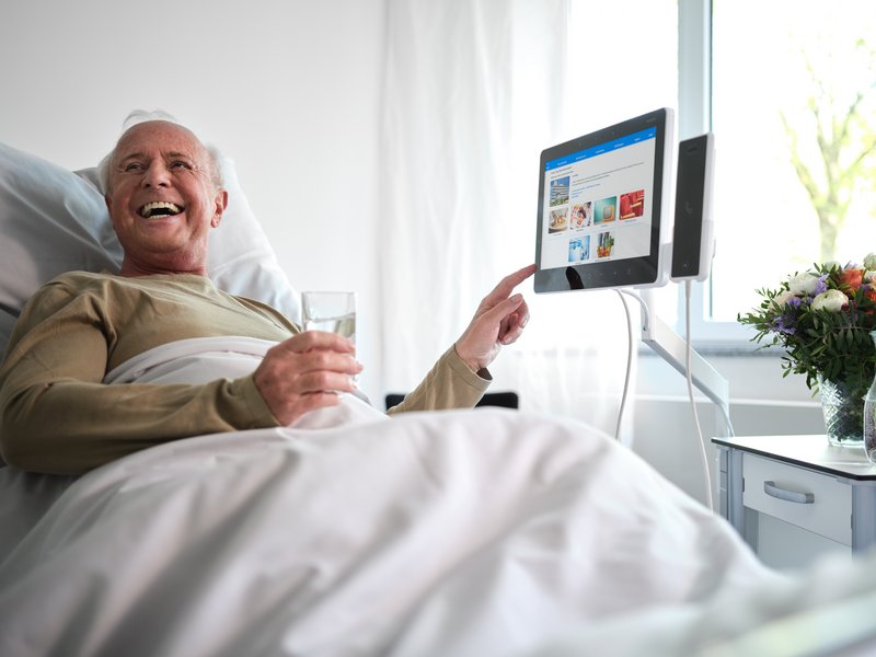 Älterer Patient im Krankenhausbett bedient lachend das BEWATEC Tablet und hält ein Glas Wasser in der Hand