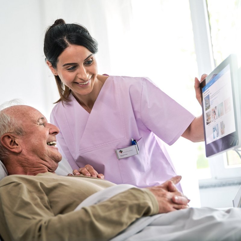 Patientenkommunikation: Junge Pflegerin und älterer Patient schauen fröhlich auf Bedside Terminal