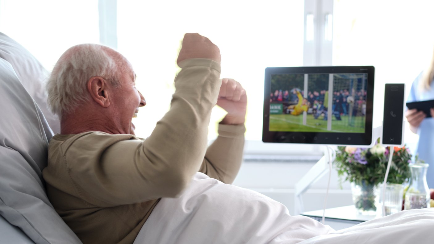 Patientenentertainment: Älterer Patient sitzt jubelnd im Krankenhausbett und schaut Fußball auf Bedside Terminal