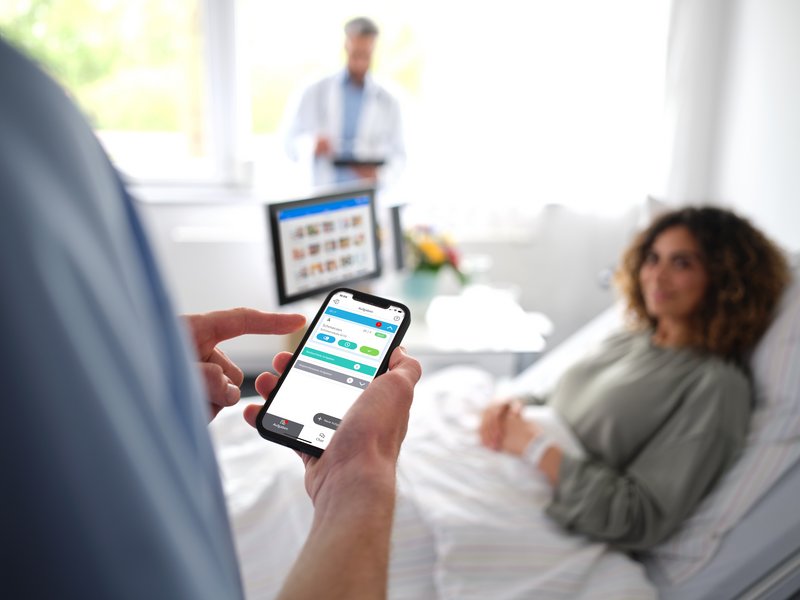 BEWATEC Patientenkommunikation | Patientin liegt im Bett und schaut zu Pfleger, der auf sein mit Smartphone mit BEWATEC.ConnectedCare schaut, Arzt mit Tablet im Hintergrund