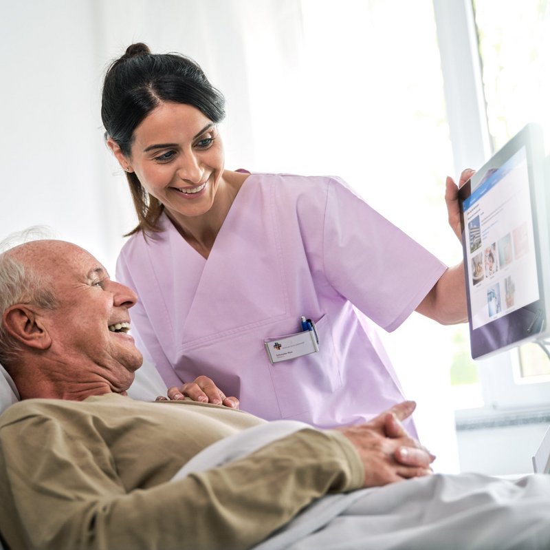 BEWATEC Patientenkommunikation | Pflegerin und Patient schauen lachend auf Bedside Terminal
