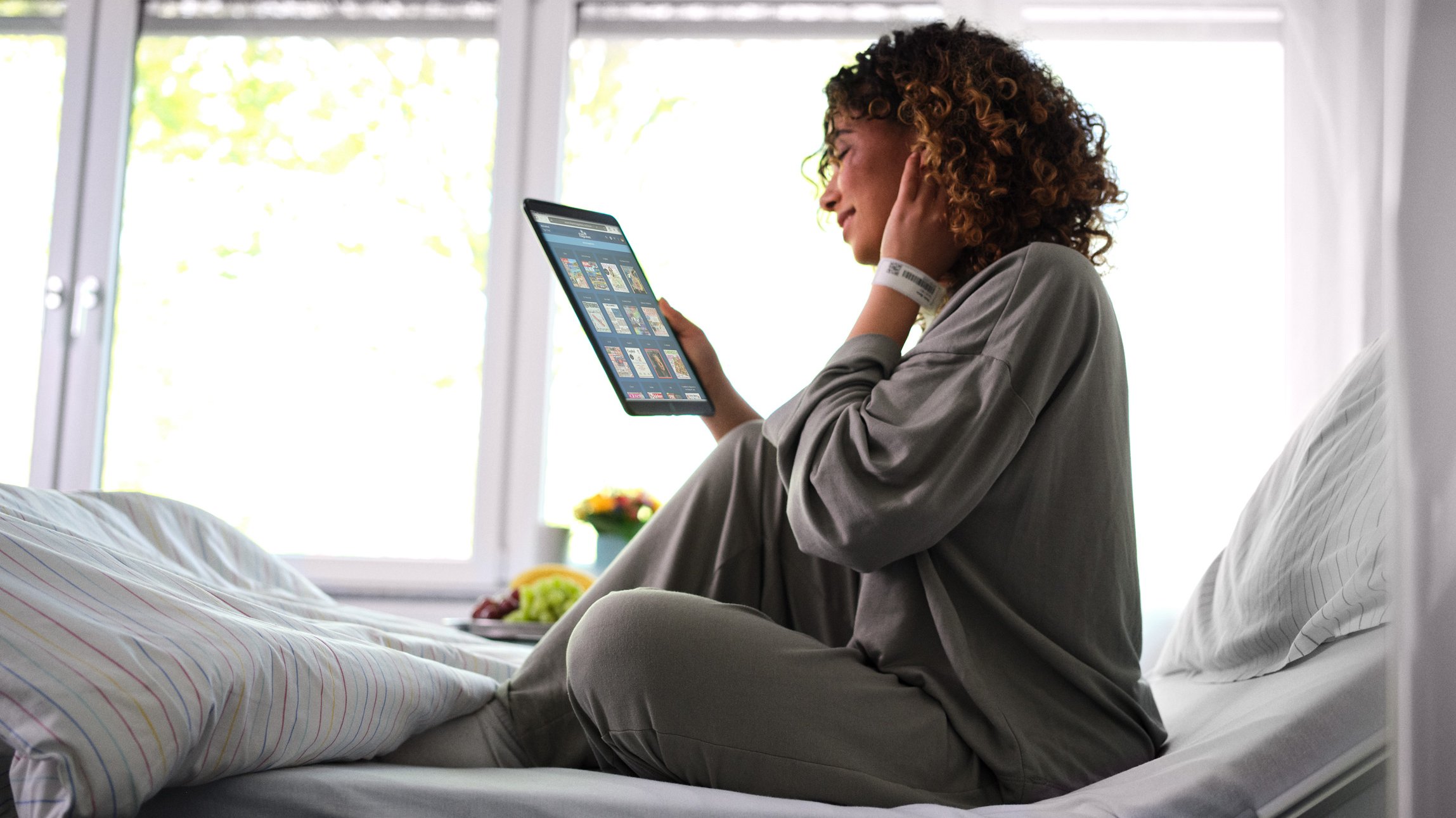 BEWATEC Patientenentertainment | Patientin sitzt im Bett und liest eMagazine auf Tablet