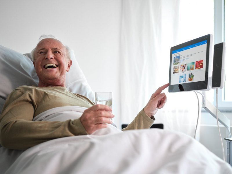 Älterer Patient im Krankenhausbett bedient lachend das BEWATEC Tablet und hält ein Glas Wasser in der Hand