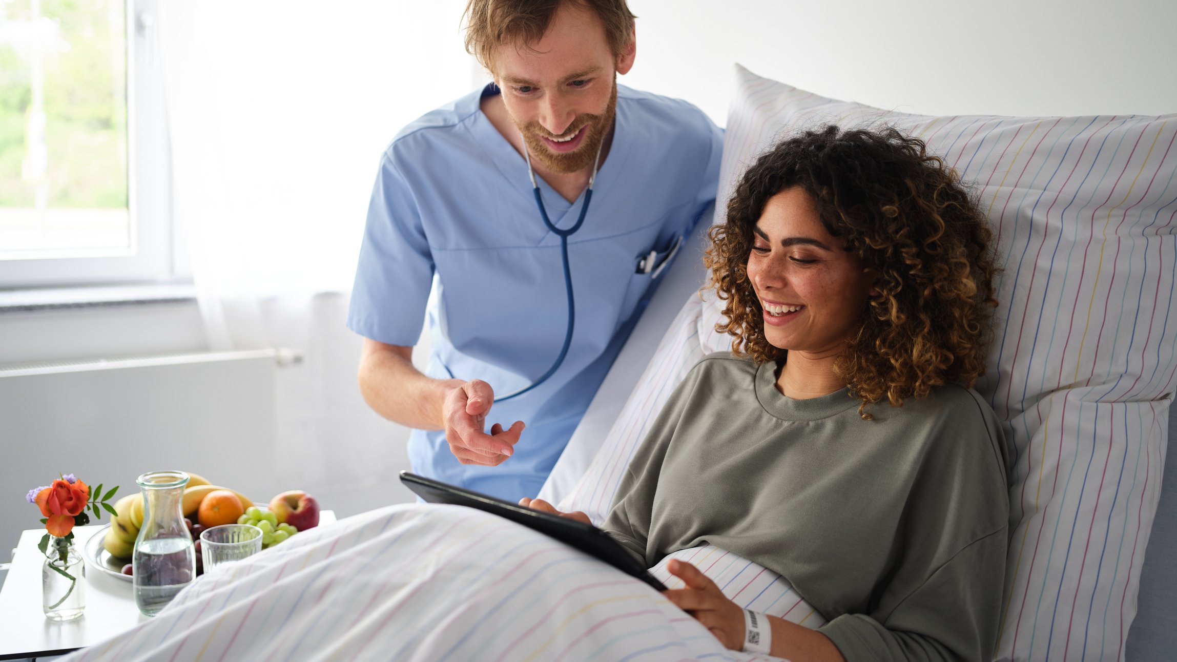 Patient Engagement | Pfleger zeigt Patientin im Krankenhausbett, wie sie auf dem iPad eine Patientenumfrage ausfüllt