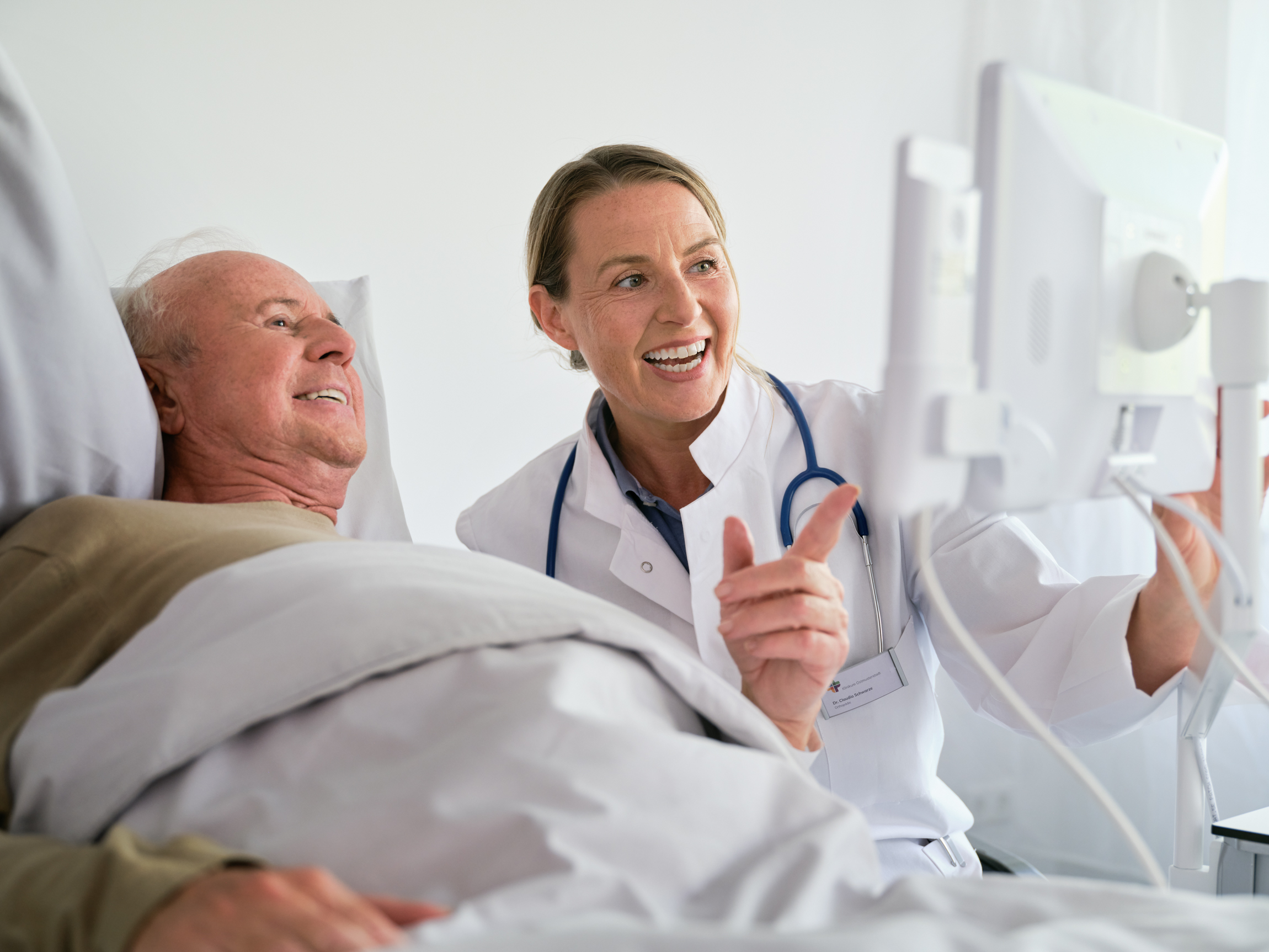 BEWATEC Digitale Kommunikation | Ärztin und Patient schauen auf Tablet am Patientenbett und lachen